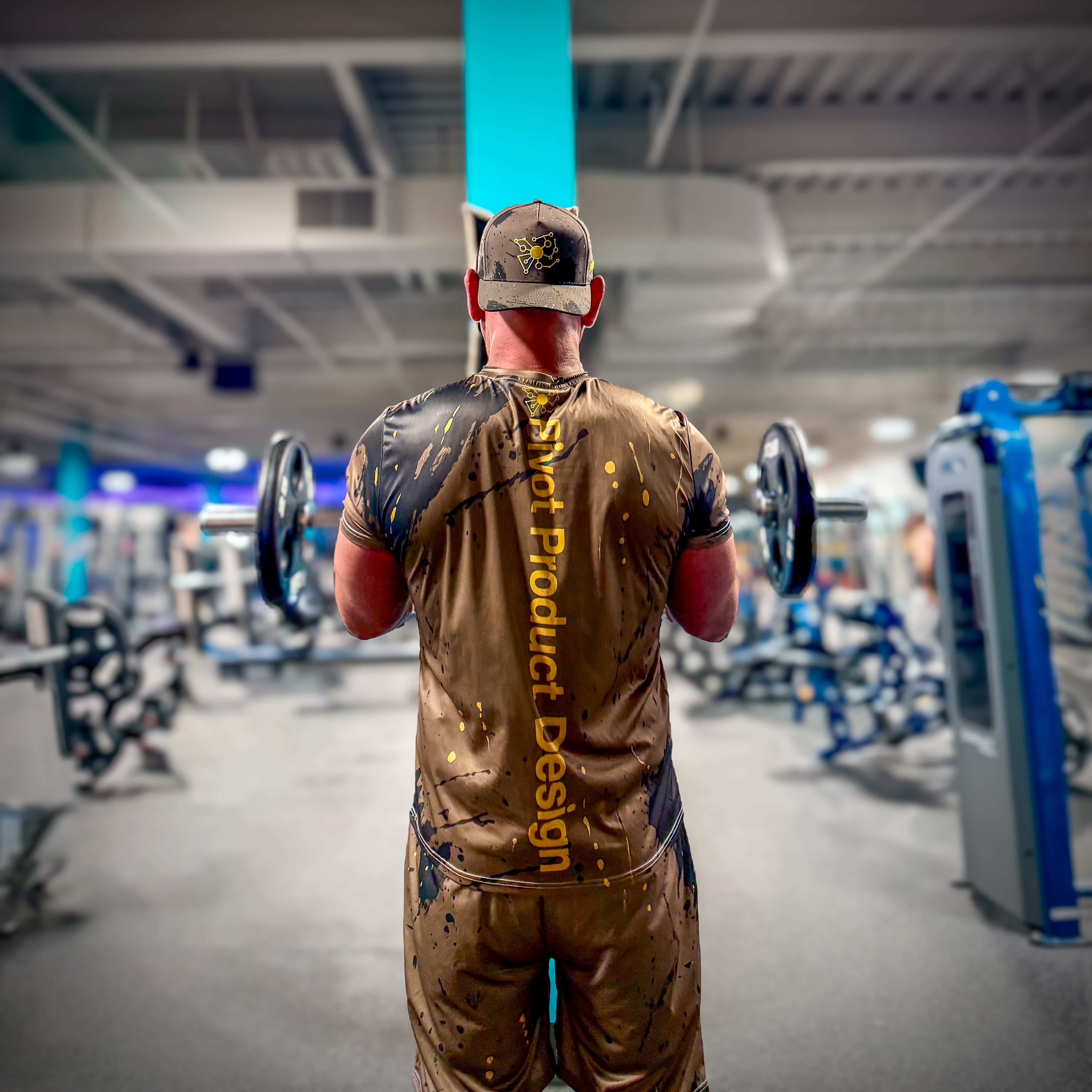 Man doing bicep curl facing media towel in gym wearing a camo outfit with 'Pivot Product Designs' text on shirt.