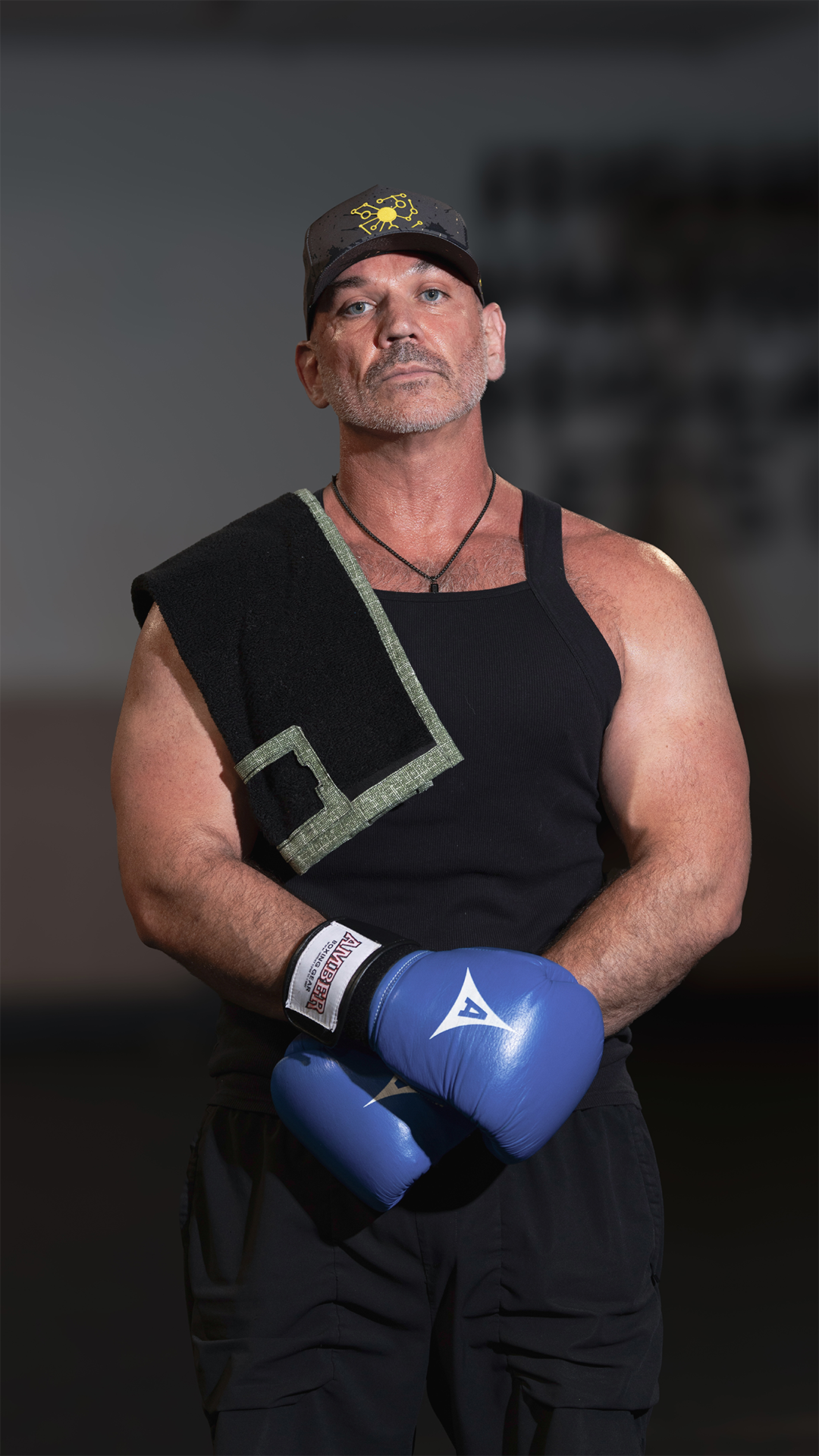 Michael McGowan, founder of PPD, wearing a black workout clothes, blue boxing gloves, and a cap with yellow pivot logo, media towel draped over shoulder..