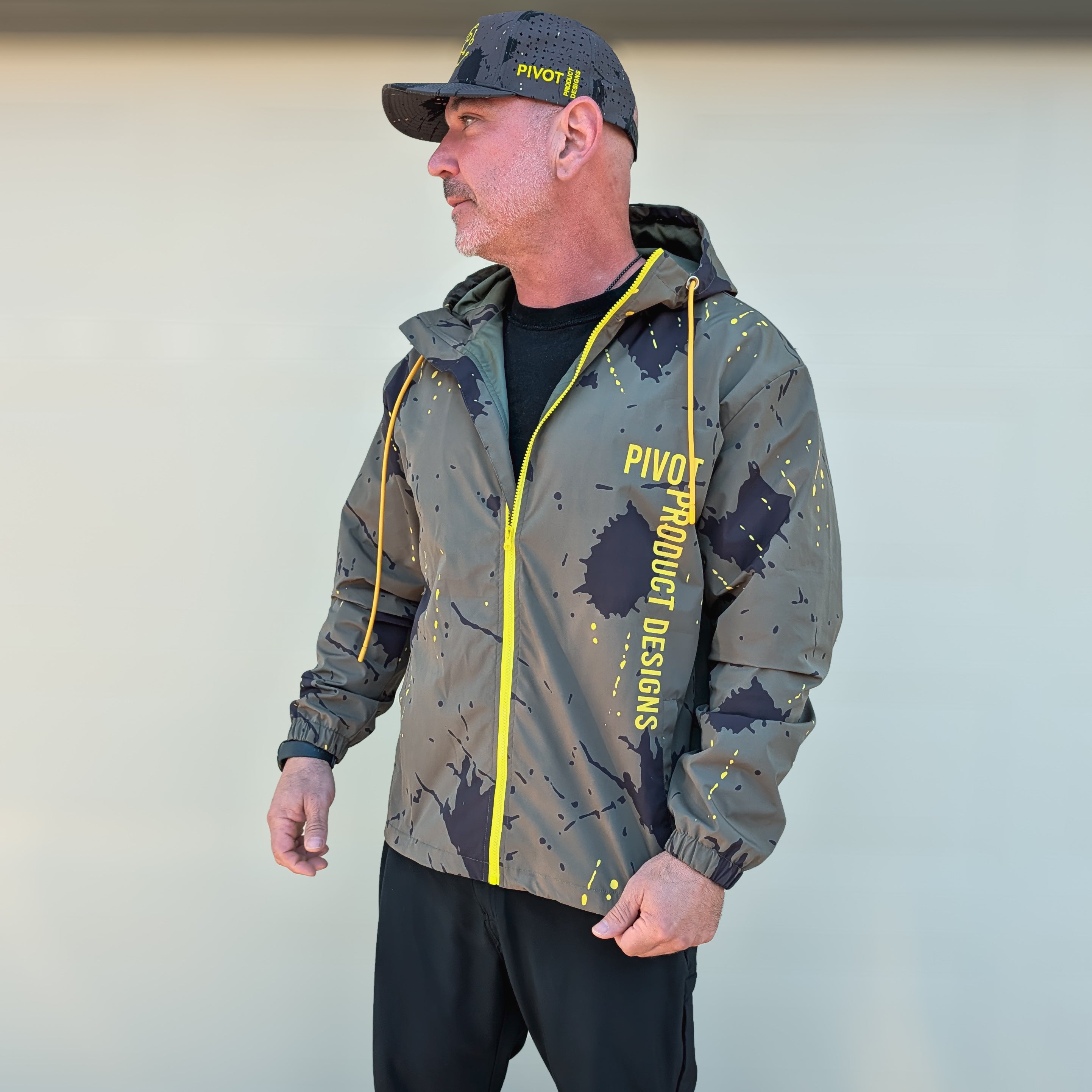 Man wearing a camouflage jacket with yellow accents and a cap, standing outside.