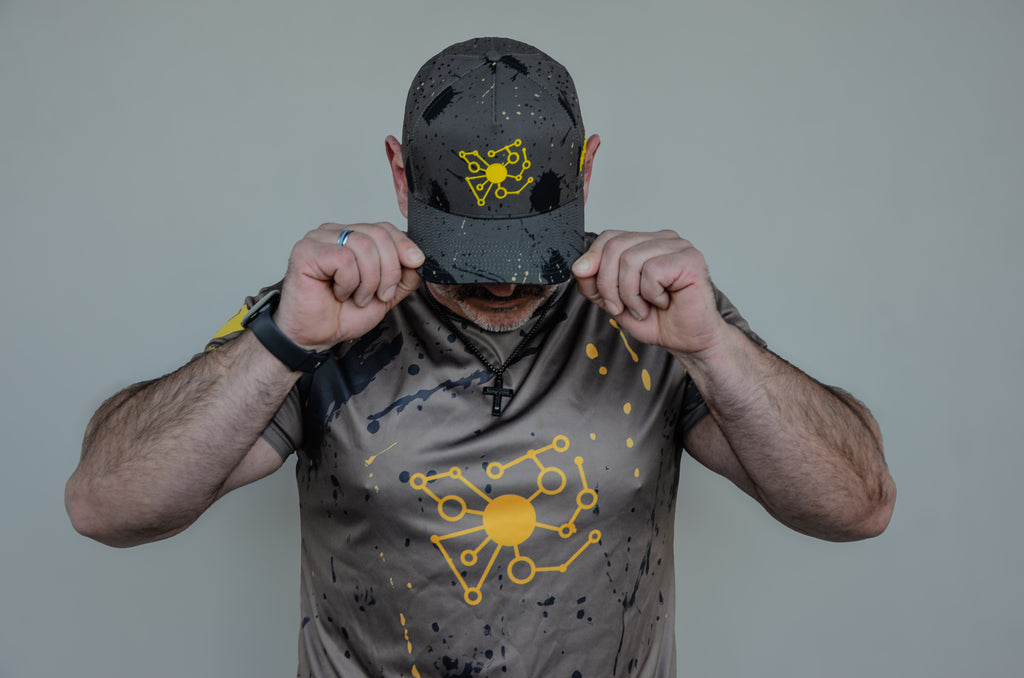 Person wearing a cap and t-shirt with Pivot Product Designs yellow logo on a camouflage background.