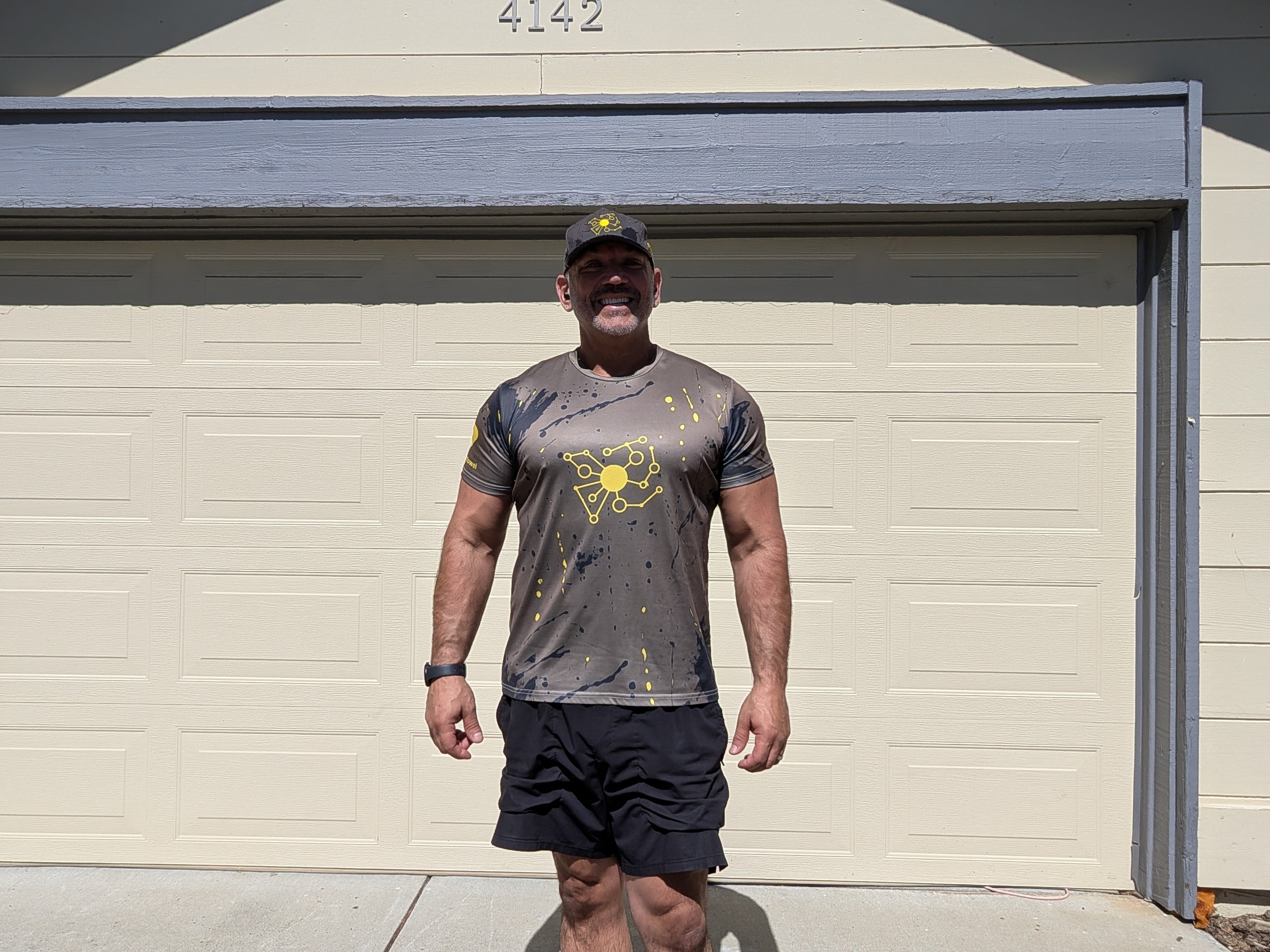 Big Mike wearing the Pivot Performance Tee — charcoal black with yellow splatter print and embroidered yellow molecular Pivot logo, front view at the garage