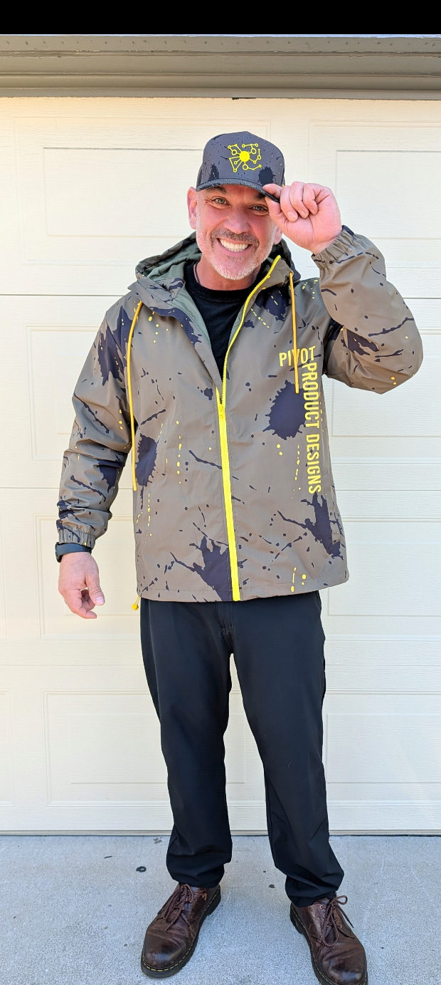 Big Mike wearing the Pivot Storm Jacket Founder Edition — olive technical shell with yellow splatter print and yellow zipper, front view at the garage