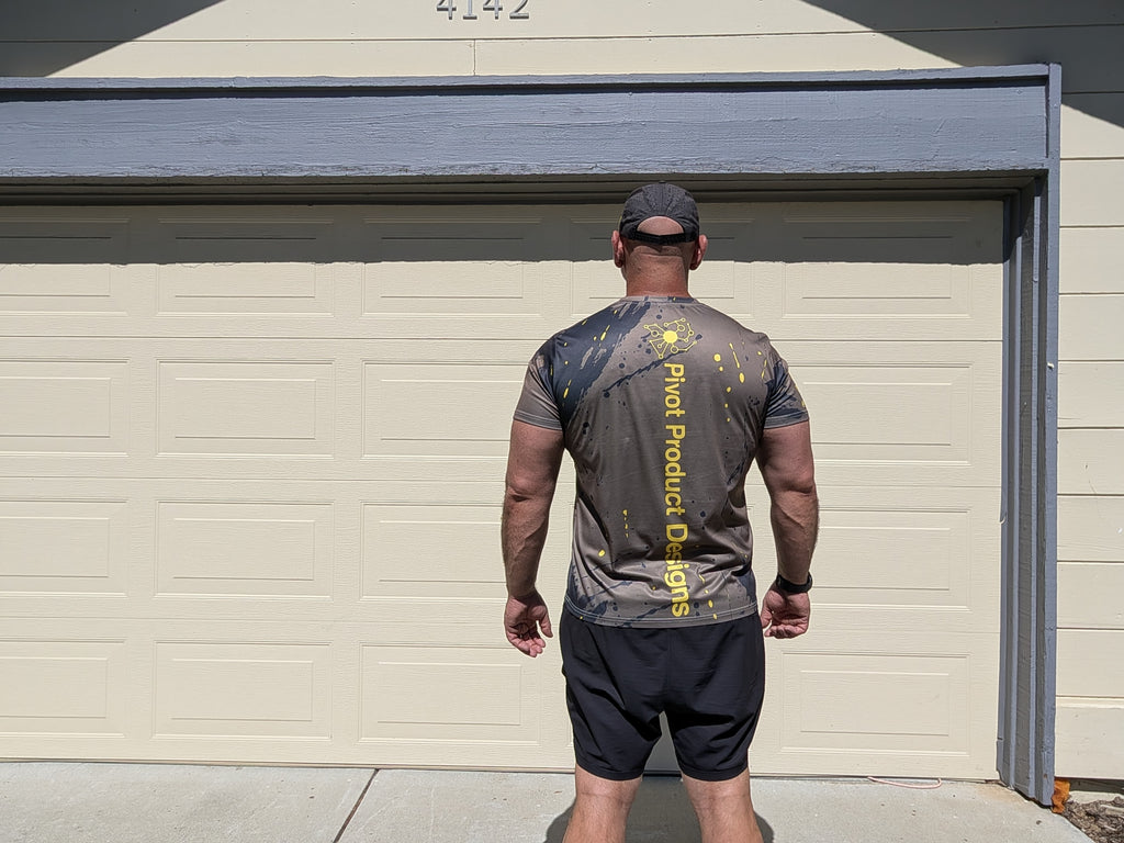 Back view of the Pivot Performance Tee showing the PIVOT PRODUCT DESIGNS wordmark and yellow splatter print on charcoal black fabric